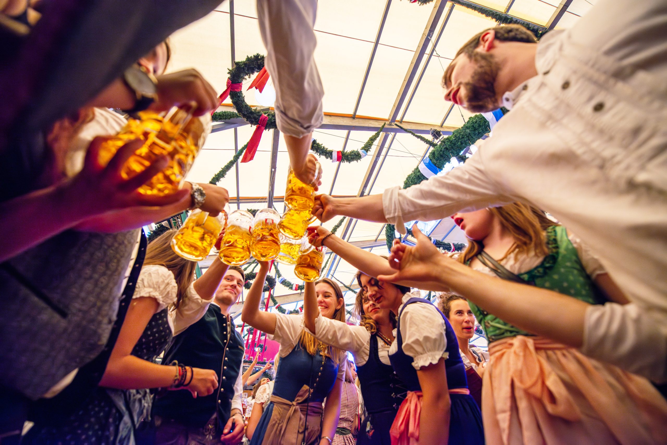 Grab the gemütlichkeit. Group of people enjoying Beer Fest tradition while toasting beer mugs in a festive tent. Celebrating friendship and German customs, they are dressed in traditional Bavarian clothing, with a joyful and communal atmosphere in the air.